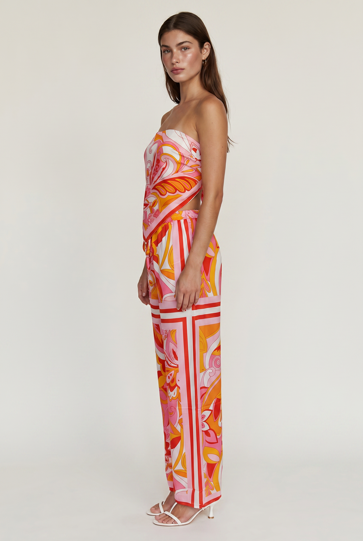 Image shoes a girl standing facing the side wearing a printed strapless top and matching pants with white heels looking at the camera.