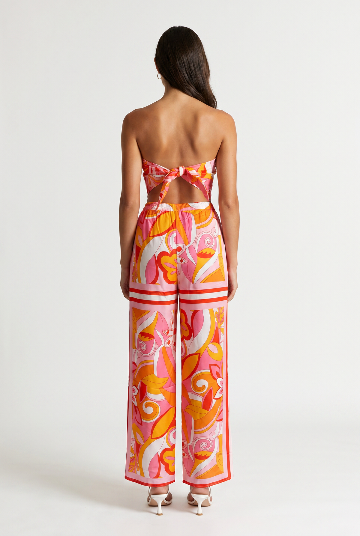 Image shoes a girl standing straight facing the back wearing a printed tied strapless top and matching pants with white heels.