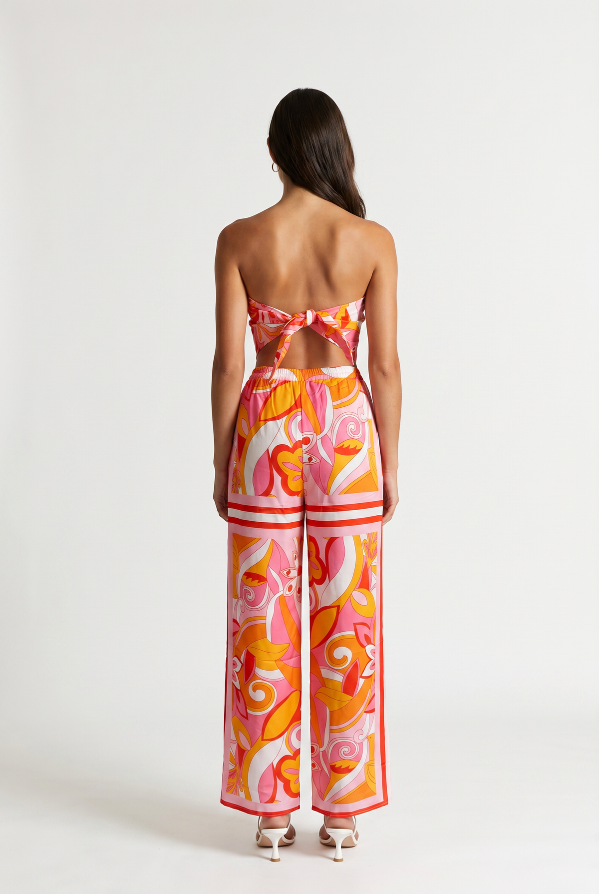 Image shoes a girl standing straight facing the back wearing a printed strapless tied top and matching pants with white heels.