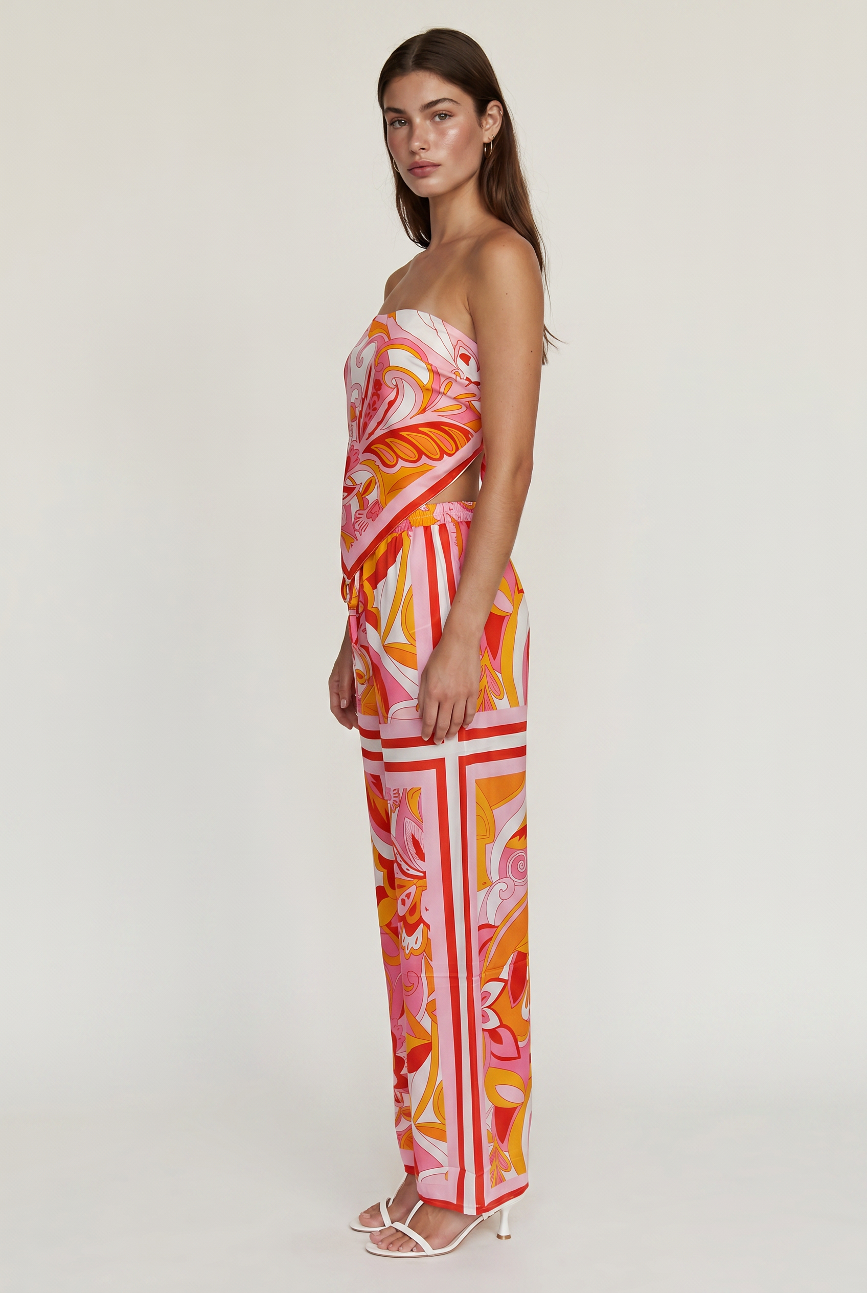 [orange] Image shoes a girl standing straight to the side wearing a printed strapless top and matching pants with white heels.