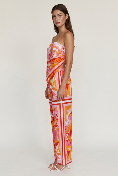 
Image shoes a girl standing facing the side wearing a printed strapless top and matching pants with white heels looking at the camera.