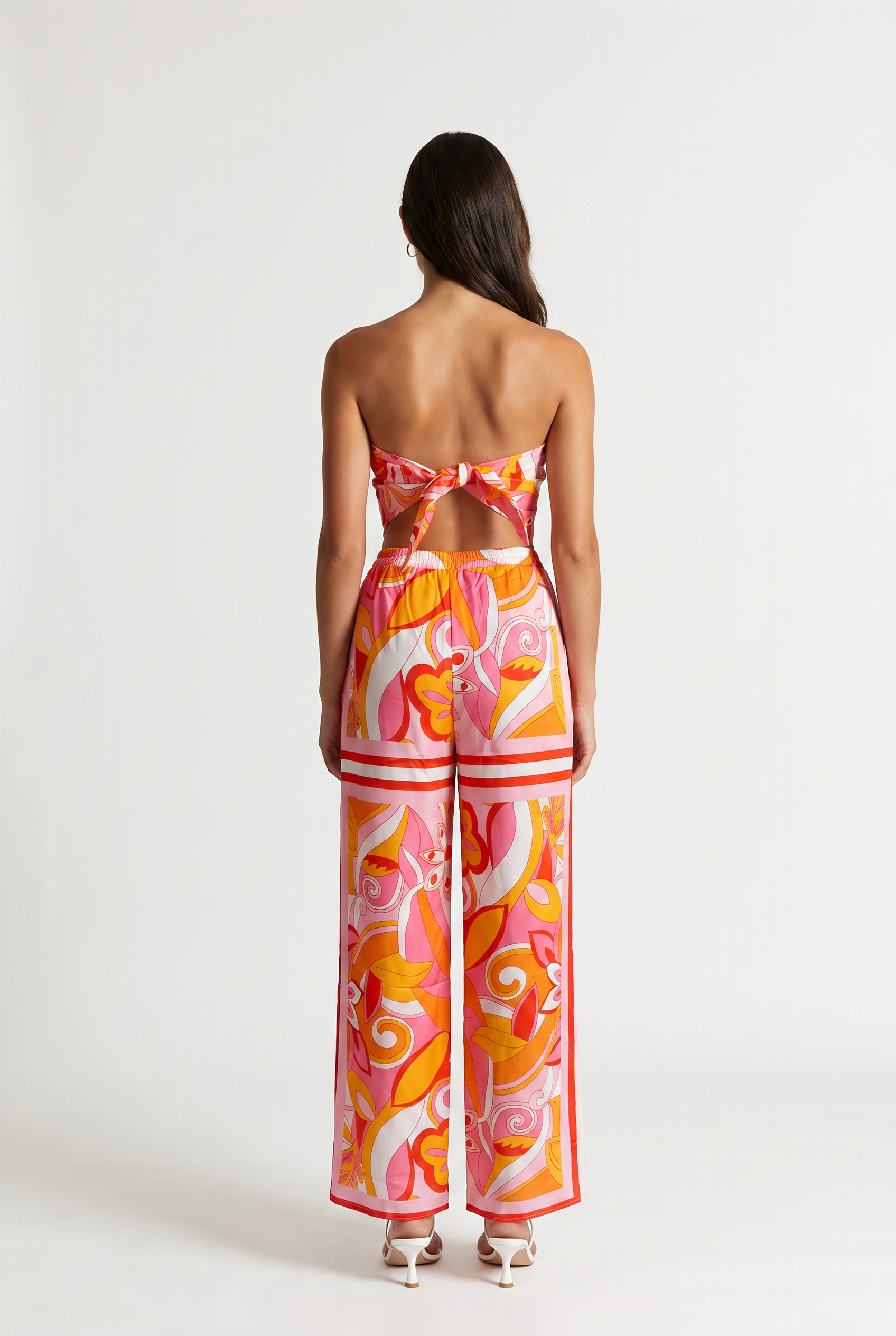 [orange] Image shoes a girl standing straight facing the back wearing a printed strapless tied top and matching pants with white heels.